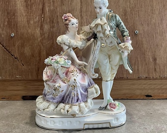May include: A porcelain figurine of a couple in 18th-century attire. The woman is wearing a pink and purple dress with gold accents and a basket of flowers. The man is wearing a green and white suit with gold accents. The figurine is on a white base.