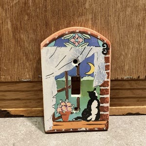 Könnte beinhalten: Dekorative Lichtschalterabdeckung aus Keramik mit einer skurrilen Szene. Das Design umfasst ein Fenster mit einer Katze, einem Mond und Blumen. Die Abdeckung hat einen Ziegelrand und ein Blumendesign oben.