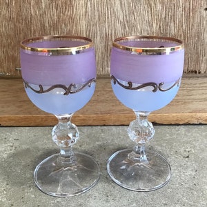 May include: Two frosted glass wine glasses with gold rims and a silver design. The glasses are a light purple color.