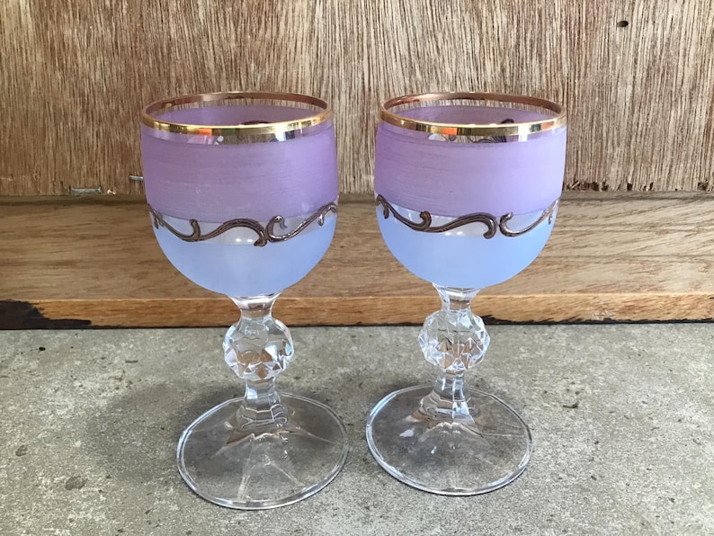 May include: Two frosted glass wine glasses with gold rims and a silver design. The glasses are a light purple color.