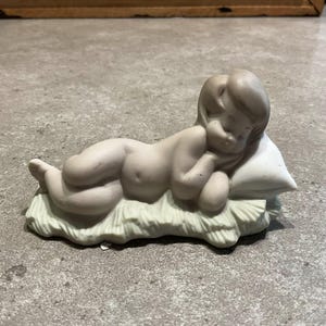Può includere: Statuina in porcellana di un cherubino addormentato. Il cherubino è sdraiato su un cuscino e un letto di paglia. La statuina è di colore beige chiaro e ha una finitura opaca. Gli occhi del cherubino sono chiusi e la sua testa è appoggiata sul braccio.