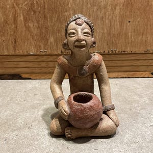 Pode incluir: Uma estatueta de terracota sentada com um rosto sorridente, segurando um pequeno pote redondo marrom-avermelhado. A figura tem um colar e pulseiras. O fundo é uma parede de madeira e uma superfície cinza.