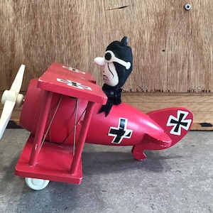 May include: A red wooden toy airplane with a black and white pilot figure. The plane has a white propeller and black crosses on the wings and tail.