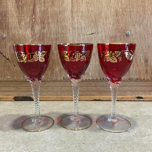 Copas de vino tinto rubí, borde dorado, tallo torcido, uvas doradas, cristalería roja