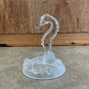 May include: A clear glass seahorse figurine with a textured surface. The seahorse is standing on a base of clear glass with a textured surface that resembles a bed of seashells.