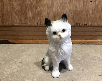 May include: A white porcelain figurine of a cat with black markings. The cat is sitting with its head tilted slightly to the side and its eyes are closed. The figurine is approximately 15 cm tall.