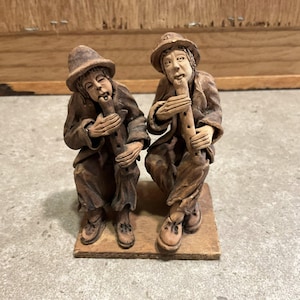 May include: A pair of brown ceramic figurines depicting musicians playing flutes. The figures are dressed in hats and coats, seated on a rectangular base. The details include facial expressions and clothing textures, creating a rustic, handcrafted appearance.