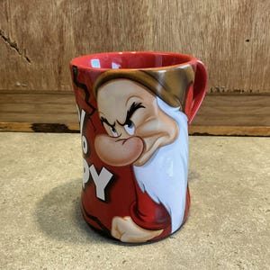 May include: Red ceramic mug featuring a grumpy-looking cartoon dwarf. The dwarf has a white beard and is wearing a red shirt. The mug reads "Grumpy" on it.