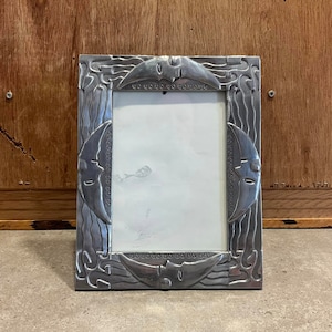 Puede incluir: Un marco de fotos plateado con un diseño ornamentado que presenta olas estilizadas y lunas crecientes. El marco es rectangular y tiene un acabado liso y metálico.