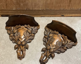 May include: Two ornate, wooden wall shelves with a dark brown finish. Each shelf features a carved design of pinecones, leaves, and a central shield-like element. The shelves are likely for decorative purposes.