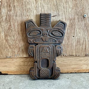 May include: A dark brown wooden carving of a stylised animal face with geometric patterns. The carving is in the style of Northwest Coast Indigenous art.