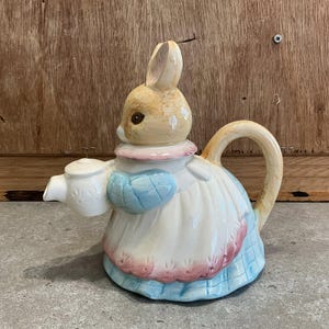 Puede incluir: Una tetera de cerámica con forma de conejito que lleva un vestido. La tetera es principalmente blanca con detalles azules y rosas. El conejito tiene pelaje marrón claro y ojos marrones. El asa y el pico son beige.