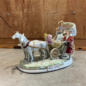 May include: A porcelain figurine of a horse-drawn carriage with two white horses, a woman in a blue dress and a man in a red coat. The carriage is decorated with yellow and pink flowers. The figurine is on a white base with green and gold accents.