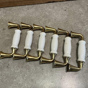 Könnte beinhalten: Sechs goldfarbene und weiße Keramik-Schranktürgriffe. Die Griffe haben eine gebogene L-Form mit einem weißen Keramikgriff und einer goldenen Metallbasis.
