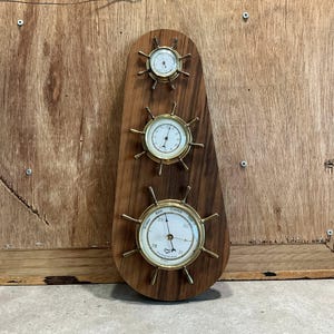 May include: A wooden weather station with three gauges. The gauges are round with brass-colored rims and hands, resembling ship's wheels. The wooden base is a dark brown color. The gauges display temperature and weather conditions.
