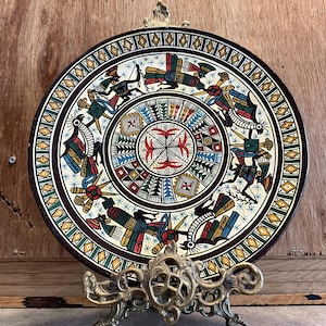 May include: A round ceramic plate with a decorative design featuring stylized figures and geometric patterns in shades of red, green, blue, yellow, and black. The plate is on a gold metal stand.