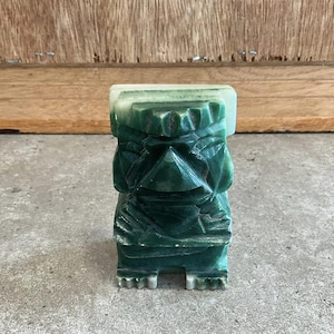 May include: A green jade carved tiki statue with a detailed face and a white top.