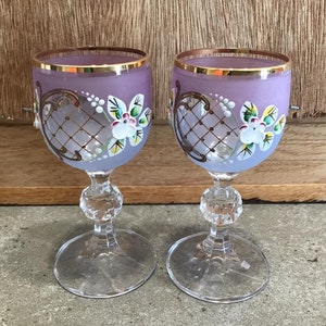 May include: Two purple glass goblets with gold rims and painted floral designs. The goblets have a lattice pattern and are frosted.