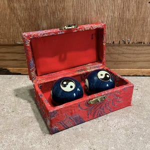 Peut inclure: Deux sphères bleues avec des symboles yin-yang blancs et noirs sont placées dans une boîte rouge à motifs avec un fermoir en laiton. La boîte est ouverte, révélant un intérieur rouge.