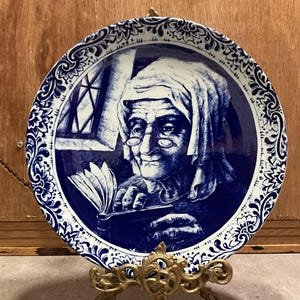 May include: A blue and white decorative plate with a detailed illustration of an elderly person reading a book. The plate has a floral border and is displayed on a gold-coloured stand.