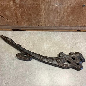 May include: A dark brown wooden carving of a stylized figure with intricate details. The carving is shaped like a long, curved blade with a pointed tip.