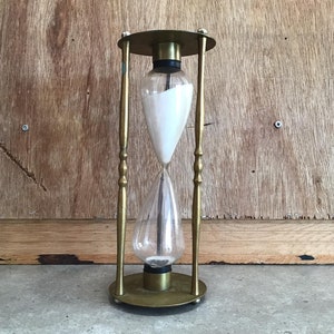 May include: An hourglass with a brass frame and base, holding white sand within two clear glass bulbs. The hourglass is supported by four brass rods and stands on a circular base. The background is a wooden surface.