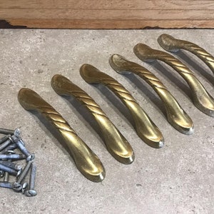 Op de afbeelding: Zes gouden kastladegrepen met een touwachtig ontwerp. De grepen zijn in een rij op een lichtgrijs oppervlak gerangschikt.