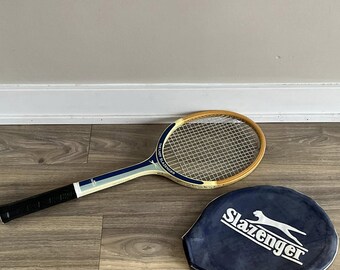 May include: A vintage Slazenger tennis racket with a wooden frame and a blue and white grip. The racket is accompanied by a blue Slazenger tennis racket cover with a white logo.