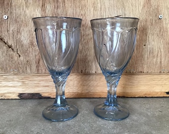 Può includere: Due calici da vino vintage azzurri. I bicchieri hanno un gambo decorativo a spirale e un design dettagliato della coppa. I bicchieri sono in vetro e sono esposti su uno sfondo di legno.