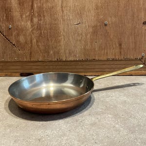 Pode incluir: Uma frigideira redonda com interior metálico polido e exterior em tons de cobre. A frigideira apresenta um cabo longo e fino de cor latão, fixado na lateral com dois rebites visíveis, estendendo-se para fora do recipiente principal.