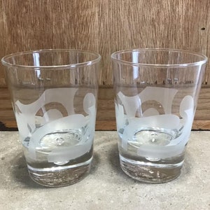 Könnte beinhalten: Zwei klare Trinkgläser mit einem mattierten Design. Die Gläser haben eine zylindrische Form und sind mit einer klaren Flüssigkeit gefüllt. Das mattierte Design scheint abstrakte Formen zu haben. Die Gläser stehen auf einer hellen Oberfläche.