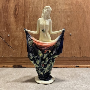 May include: A cream-colored ceramic figurine of a person holding a draped fabric that forms a vase. The fabric features a dark blue base with a pattern of green leaves, red flowers, and yellow butterflies. A small yellow butterfly is on the chest.
