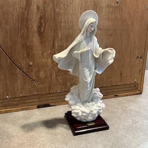 May include: A white porcelain statue of a woman with a halo, standing on a cloud-like base. The statue has flowing robes and is mounted on a dark wooden base. The background is a wooden panel.