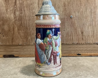 Könnte beinhalten: Ein hoher Bierkrug aus Keramik mit einem detaillierten, farbenfrohen Relief von Menschen in Trachten. Der Krug hat einen Deckel aus Zinn und einen dekorativen Sockel. Der Hintergrund ist eine Holzoberfläche.