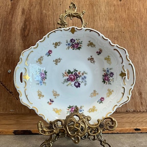 Puede incluir: Un plato de servir de porcelana blanca con ribetes dorados y diseños florales. El plato tiene dos asas y se muestra en un soporte de metal dorado.