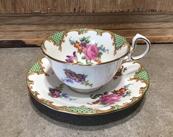 Può includere: Una tazzina da tè e un piattino bianchi con un motivo floreale verde e oro. La tazza ha un manico delicato e il piattino ha un bordo ondulato.
