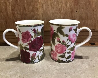 May include: Two white ceramic mugs with floral designs. Both mugs feature pink and red roses with green leaves on a white background. The mugs have gold trim around the top.
