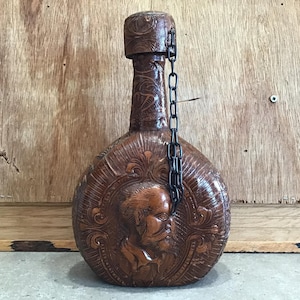May include: A brown leather bottle with intricate carvings and a metal chain. The bottle has a round body and a narrow neck. The carvings on the bottle depict a man's face and floral designs.