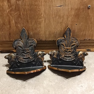 Może przedstawiać: Para vintage'owych podpór do książek Boy Scouts wykonanych z metalu. Każda podpórka zawiera brązowy emblemat Boy Scouts z lilią i napisem "Boy Scouts Be Prepared". Podpórki mają kształt bobra.