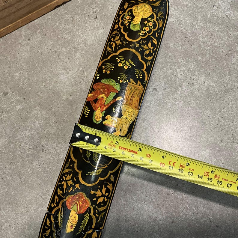 May include: A black, decorative, wooden object with ornate, hand-painted figures and floral designs. The object is approximately 17 inches long. The artwork features orange, green, and yellow accents. A yellow tape measure is placed on top of the object.