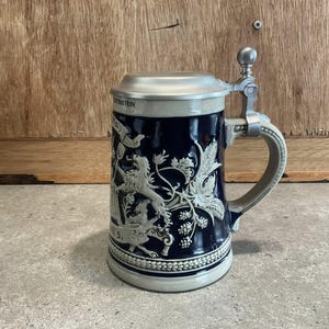 May include: A vintage beer stein with a hinged metal lid and handle. The dark blue ceramic body features a relief design of a lion, floral elements, and text. The stein has a light gray base and rim.