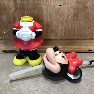 May include: A Minnie Mouse-themed tumbler with a red dress, white gloves, and yellow shoes. The tumbler is missing its head, which is lying on its side with a red bow and a straw. The tumbler is made of plastic.