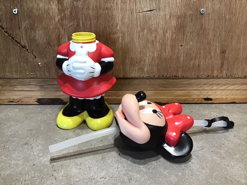 May include: A Minnie Mouse-themed tumbler with a red dress, white gloves, and yellow shoes. The tumbler is missing its head, which is lying on its side with a red bow and a straw. The tumbler is made of plastic.