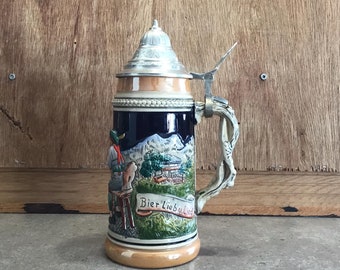 May include: A ceramic beer stein with a silver lid and handle. The stein features a colorful scene of a man in traditional clothing with a mountain backdrop. The text "Bier Lieb u Lied" is printed on the stein.