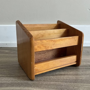 May include: A wooden desk organizer with three compartments. The organizer is made of light brown wood and has a slanted design. The compartments are separated by horizontal wooden dividers. The organizer is designed to hold mail, documents, or office supplies.