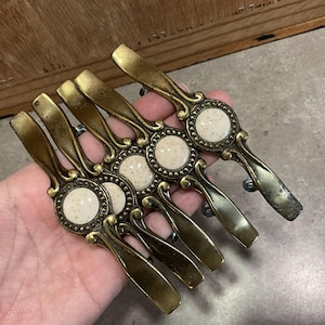 May include: Set of five antique brass cabinet pulls with a decorative design and a white ceramic center. Each pull has a round, slightly raised ceramic center with a speckled texture.