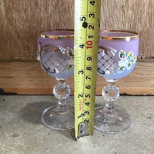 May include: Two small, clear glass shot glasses with a frosted lavender finish. Each glass has a gold rim and intricate painted floral designs in white and gold.