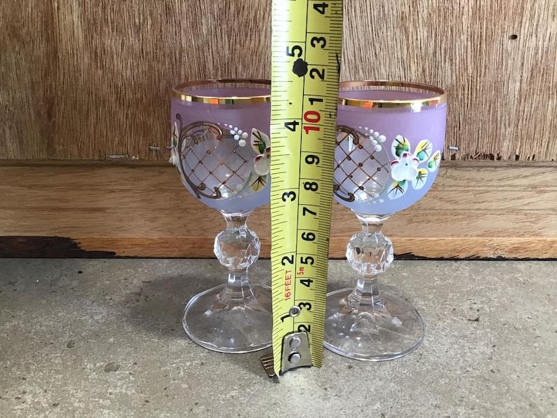 May include: Two small, clear glass shot glasses with a frosted lavender finish. Each glass has a gold rim and intricate painted floral designs in white and gold.