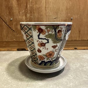 May include: A decorative ceramic planter with a matching saucer. The planter features a white background with floral and geometric patterns in shades of orange, brown, blue, and green. The planter is approximately 15 cm tall.
