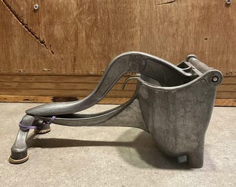 May include: A vintage, cast-aluminum citrus juicer with a lever arm and a built-in strainer. The juicer is silver-colored and has a sturdy base with rubber feet. A purple rubber band secures the lever. The design suggests it's for squeezing lemons, limes, or oranges.